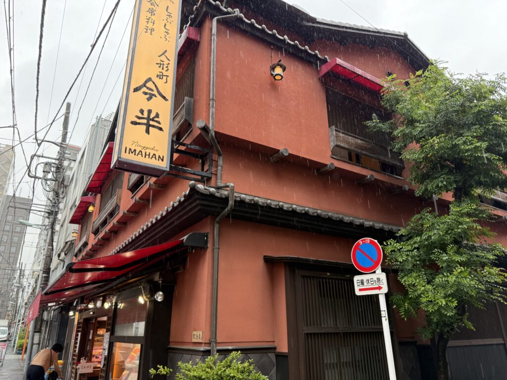 すき焼きの老舗、人形町今半本店の伝統的な日本家屋の外観。瓦屋根、白壁、木の格子、藍色の暖簾、そして店名が書かれた大きな提灯が特徴的。
