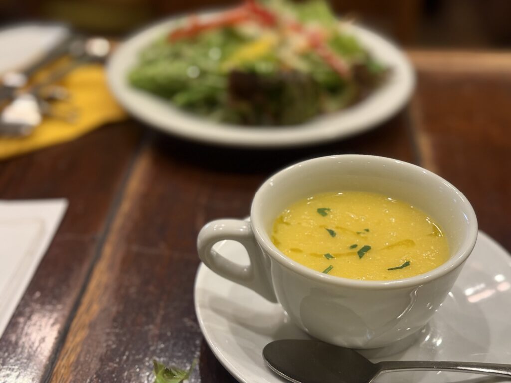 Corn soup from PIZZA DA BABBO. A yellow soup in a white deep plate, garnished with green herbs and black pepper, with a spoon.