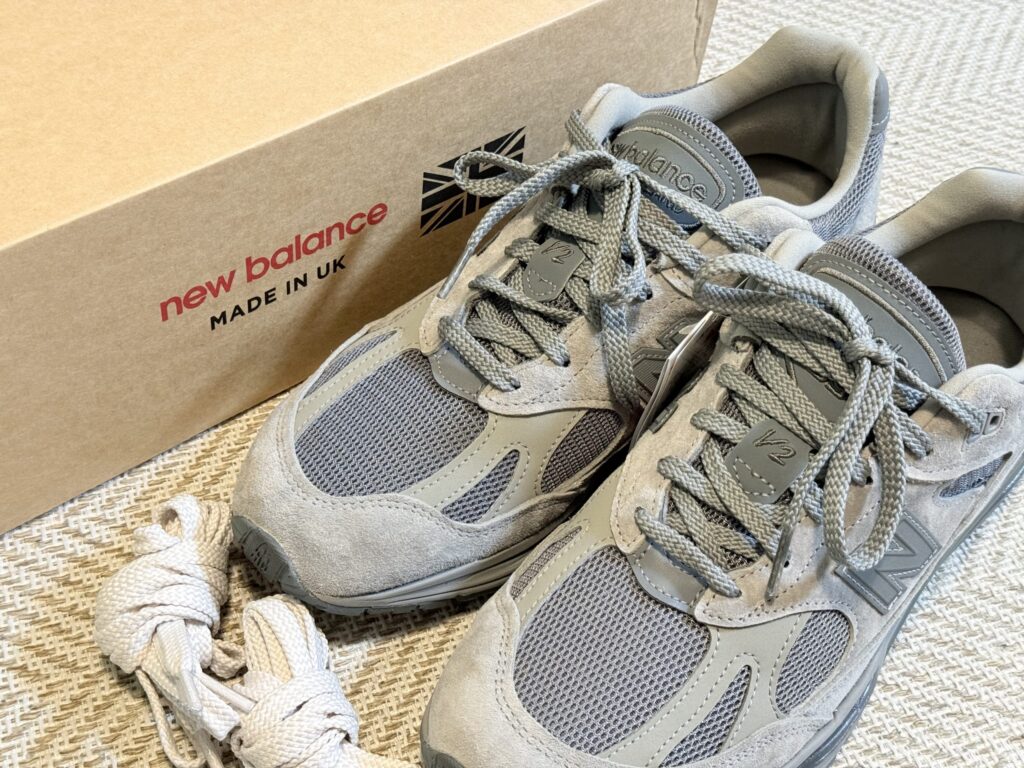 グレーとブラウンのthisisneverthat x New Balance 991v2スニーカーを、付属品(予備シューレース、タグ、シューズバッグ)と共に横から撮影したクローズアップ写真。