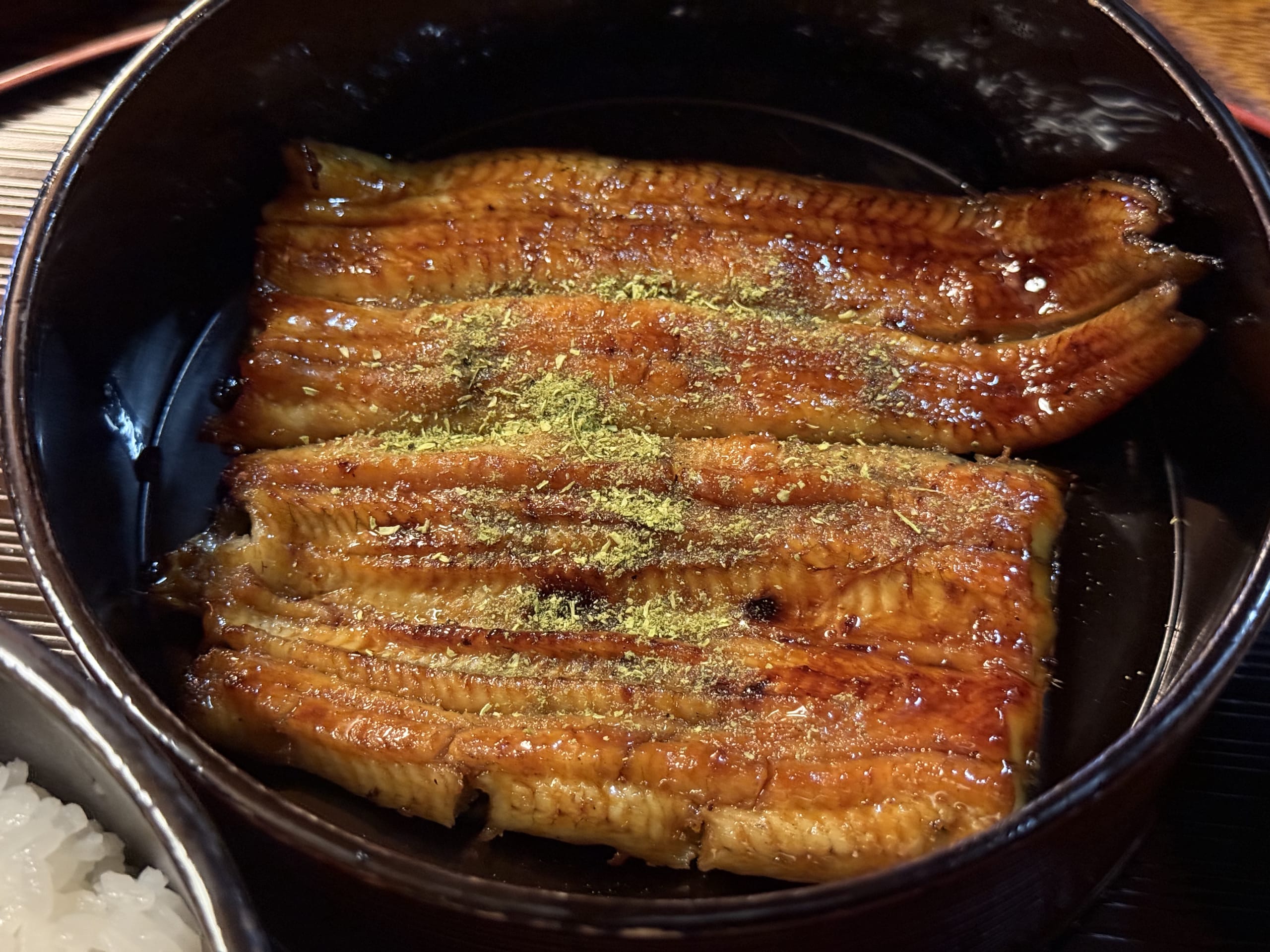 黒い器に盛られた鰻料理に、自家製の山椒が鮮やかに振りかけられているクローズアップ写真。