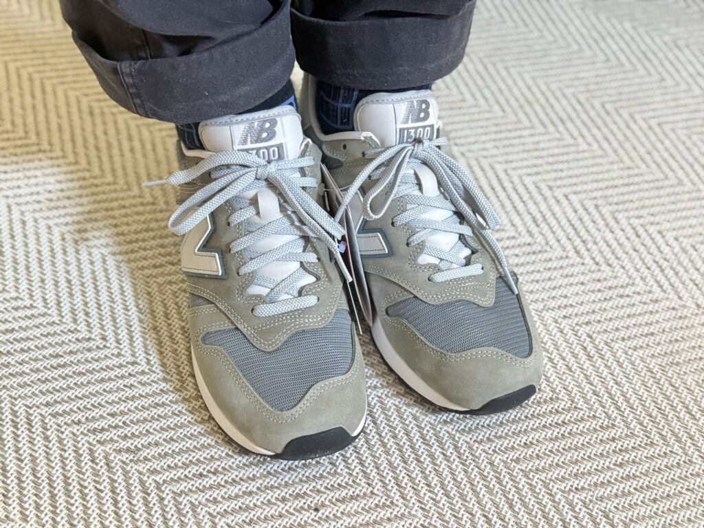 Close-up photo of feet wearing grey New Balance U1300JP sneakers, standing on asphalt.