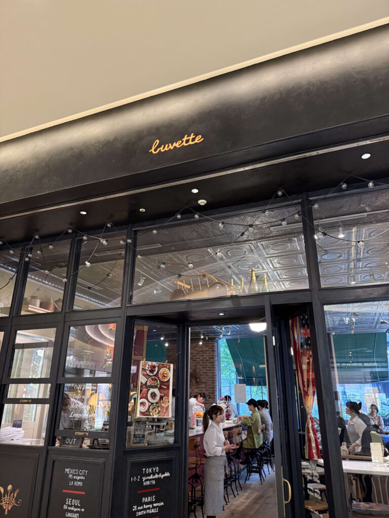 パリのカフェ「Buvette」の外観。赤い日除けと緑色の壁、店先にテーブルと椅子が置かれている様子。