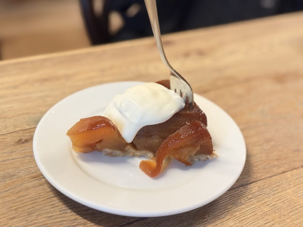 A slice of Tarte Tatin with beautifully shiny, caramel-colored apples, served on a white plate with a fork.