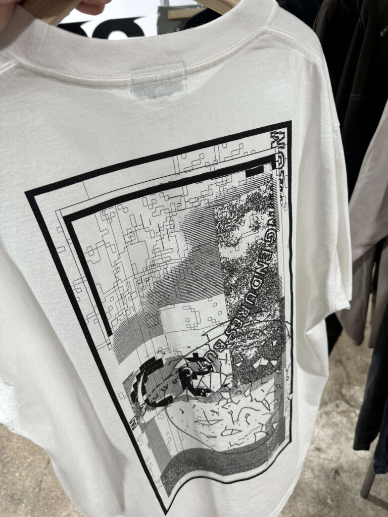 白い背景にハンガーで掛けられたC.EのTシャツ。背面に青、赤、白を中心とした複雑な幾何学的グラフィックプリントが施されている。
