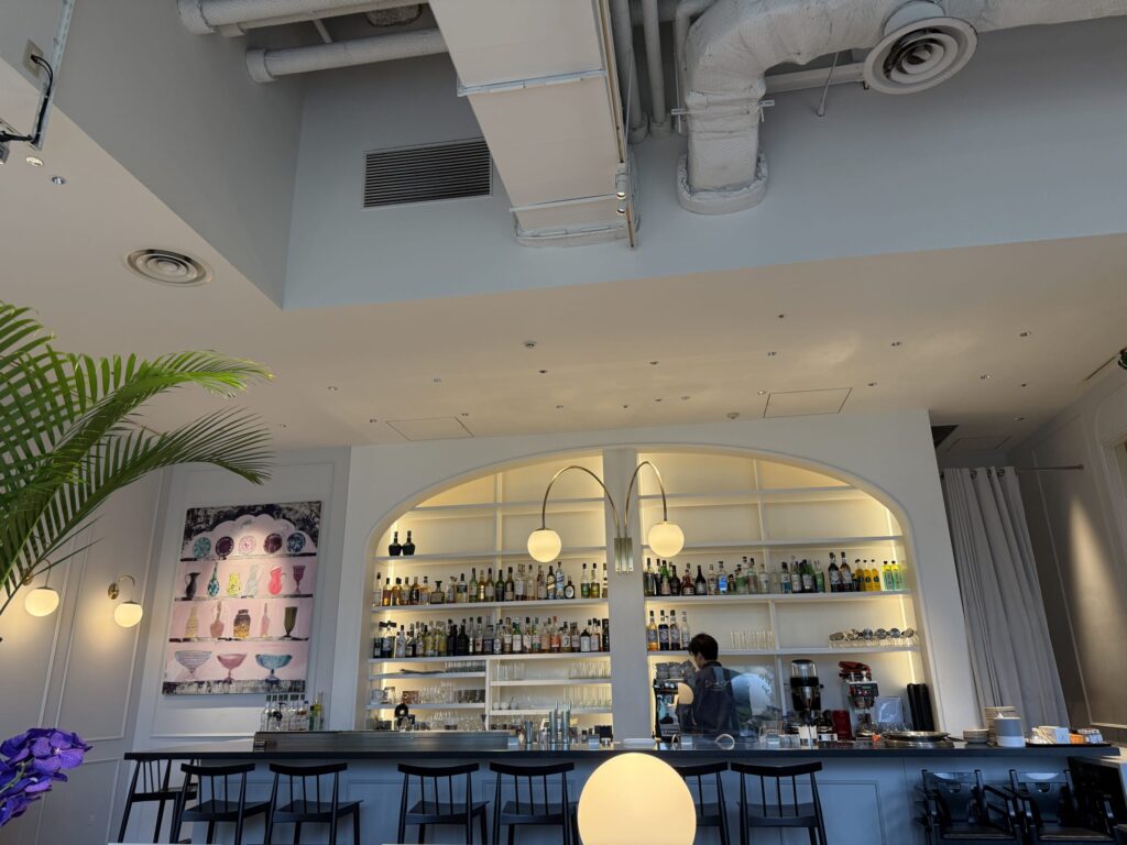 A bar corner illuminated by warm lighting, featuring a wooden counter, high stools, and a brick wall in the background with shelves of bottles.