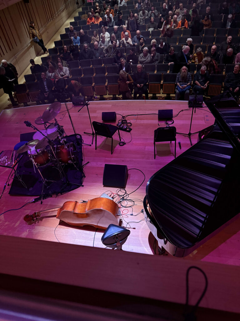 Overlooking a dimly lit stage from a balcony seat with a red railing in a dark theater. The stage is illuminated by bluish-white light.