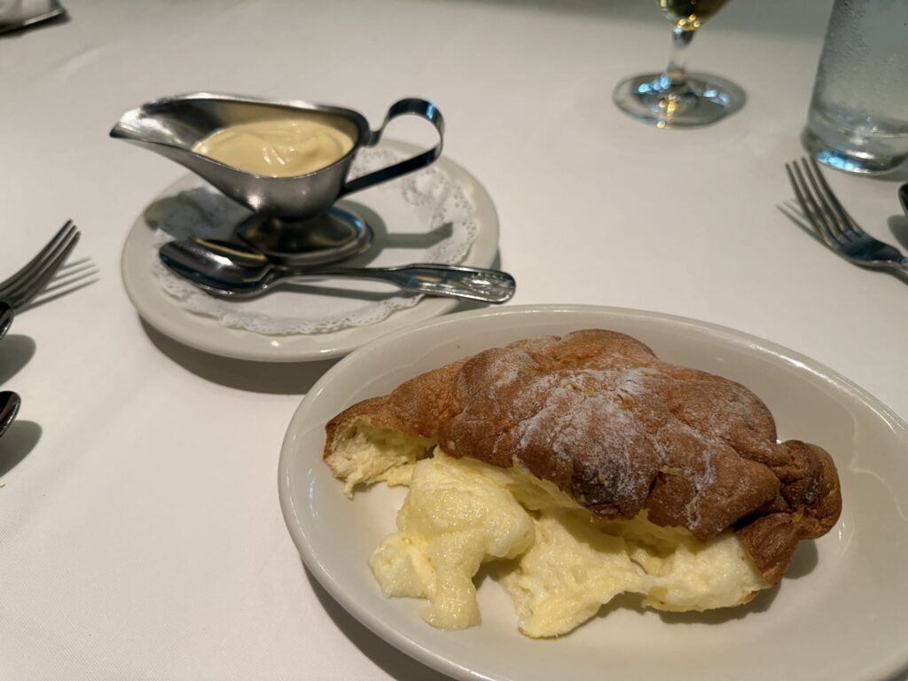 A fluffy soufflé on a white plate, with a piece cut out by a spoon, revealing its airy texture. A small pitcher with custard sauce is in the background.