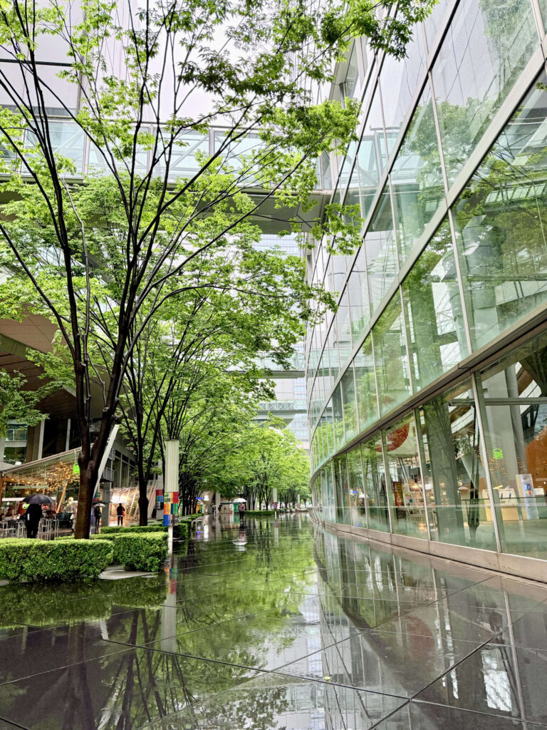 雨の日の東京国際フォーラム。二棟のビルに挟まれた通路の濡れた床が、周囲の景色や木々を鏡のように映し出している。