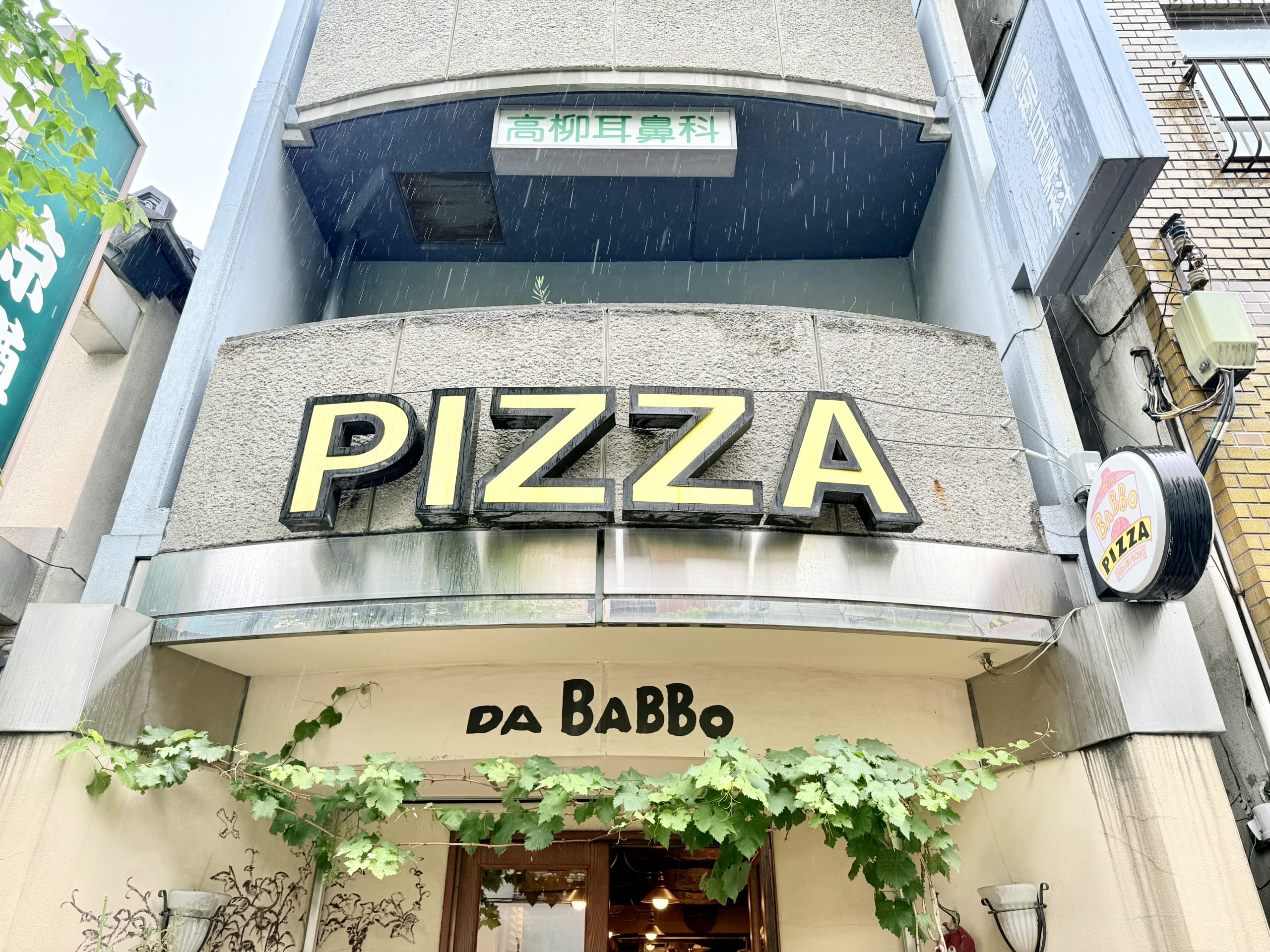 The pop-designed exterior of PIZZA DA BABBO, featuring a distinctive red door and window frames, with the store's logo and pizza illustrations on the white wall.