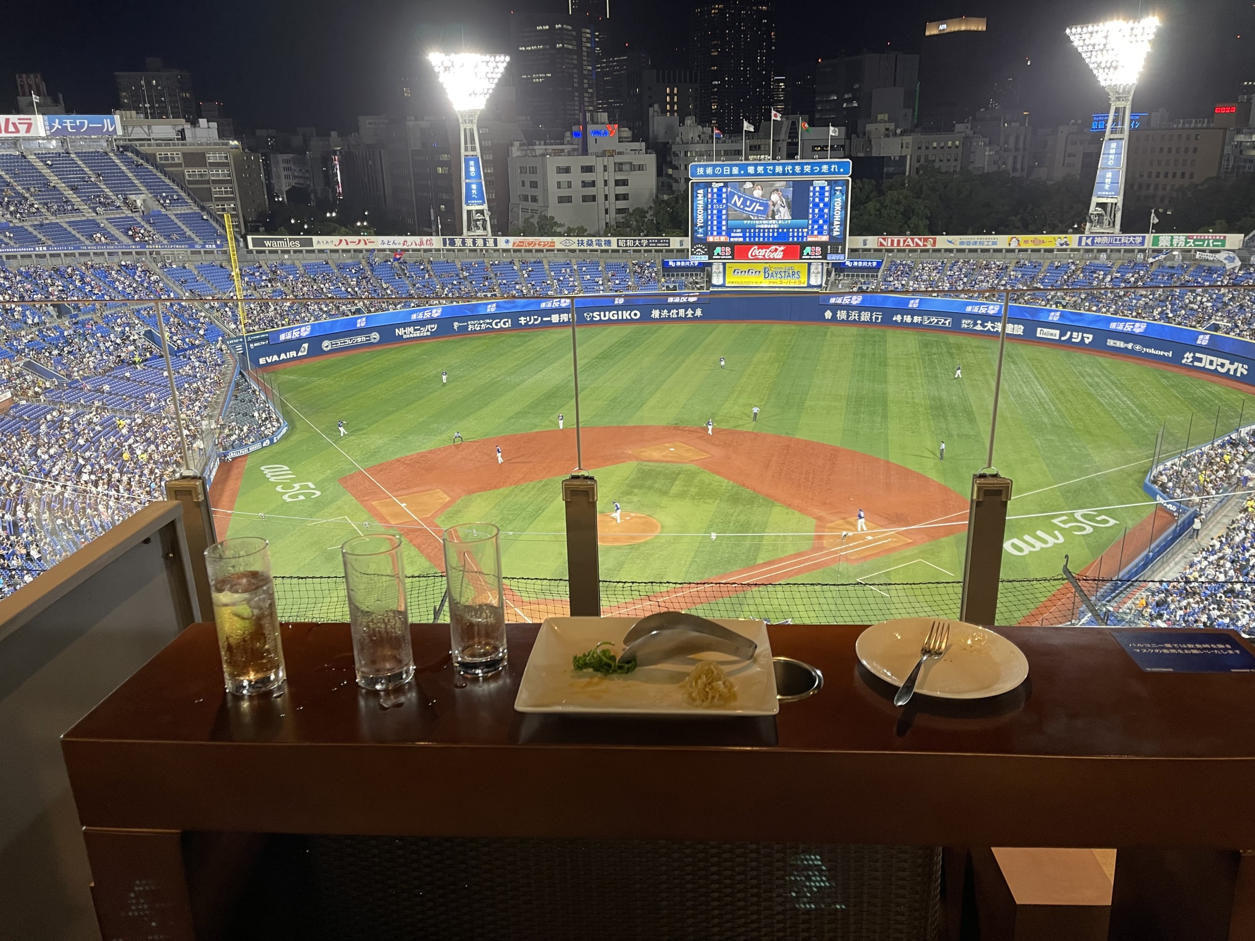 スタジアムのバルコニー席のテーブルに置かれたビールと料理。背景には試合を映す大型スクリーンがぼやけて見える。