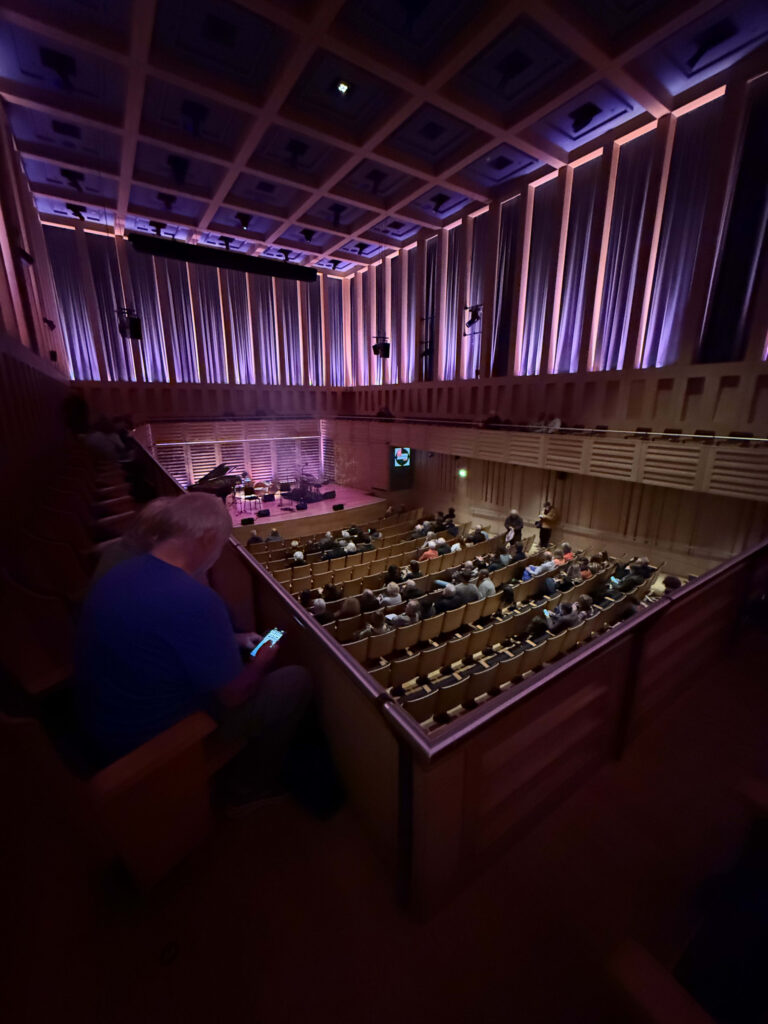 Interior of a vast concert hall, showing numerous arena seats facing the stage and multiple tiers of balcony seating.