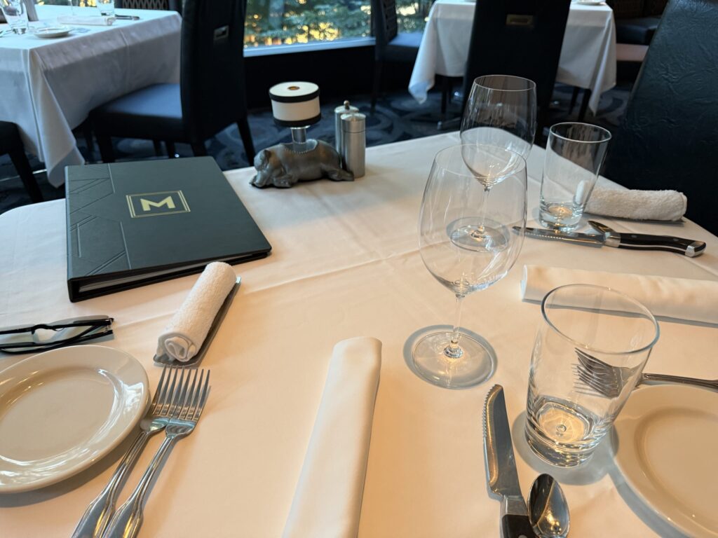 白いテーブルの上に置かれたミニマルな白い食器のセット。皿の上にはスプーンが入ったボウルが置かれ、その横にフォークとナイフが並んでいる。