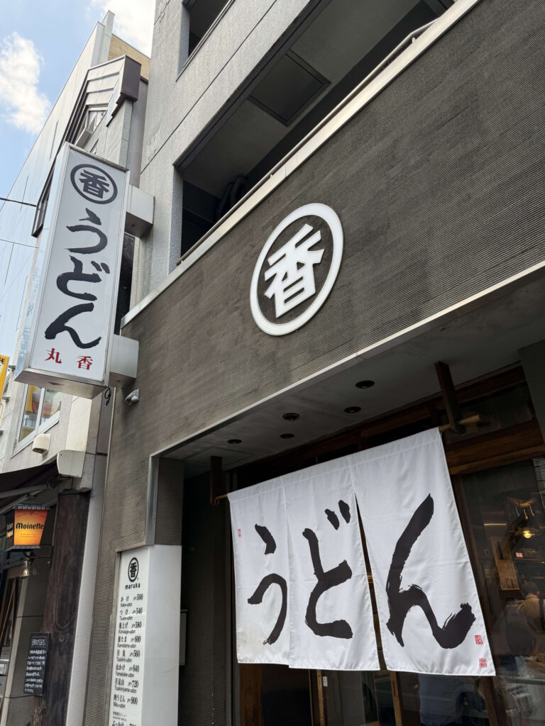 うどん屋「丸香」の店舗外観。白い暖簾に黒文字で「うどん」と書かれている。