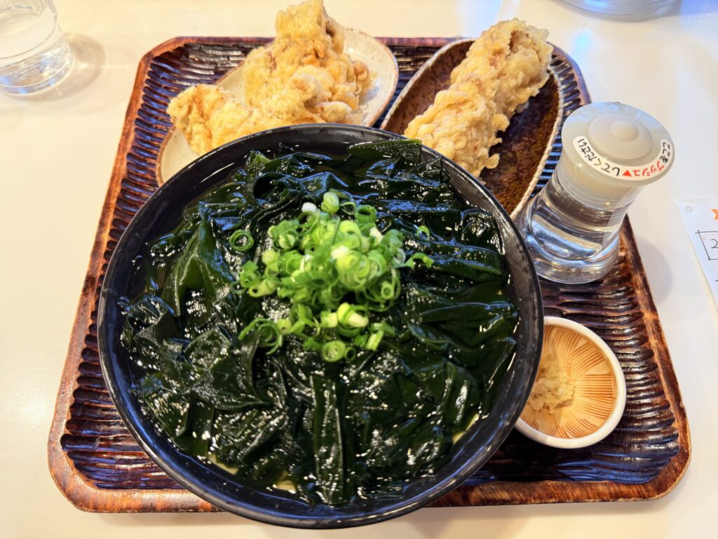 冷たいわかめうどん、かしわ天、ちくわ天、そして味変用の酢が木製のトレイに載っている様子を上から撮影した写真。