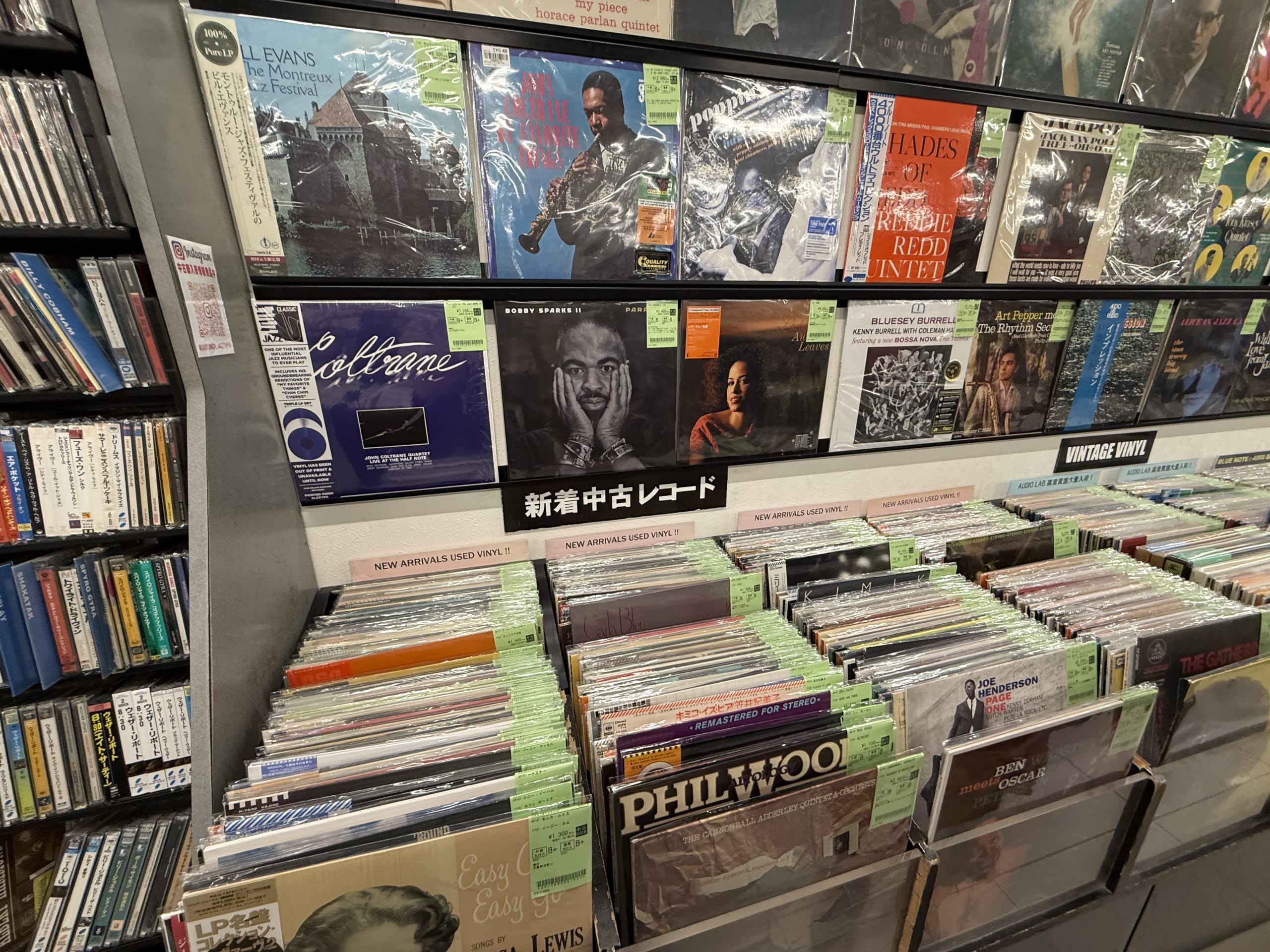 古着屋の新入荷コーナー。ハンガーに様々な衣類が掛けられ、「新入荷コーナー」と書かれた手書き風のポップが見える。