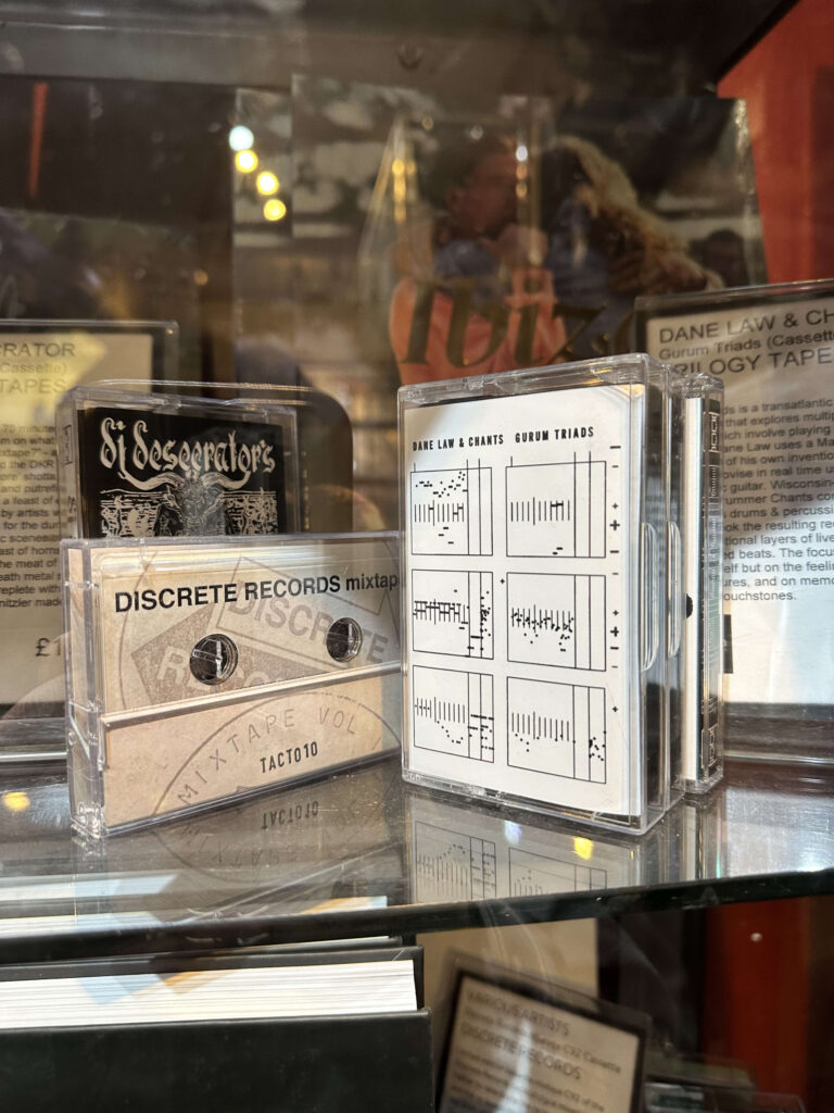 A showcase displaying cassette tapes to the right of a store entrance, with DANE LAW & CHANTS' album 'GURUM TRIADS' visible in the center.