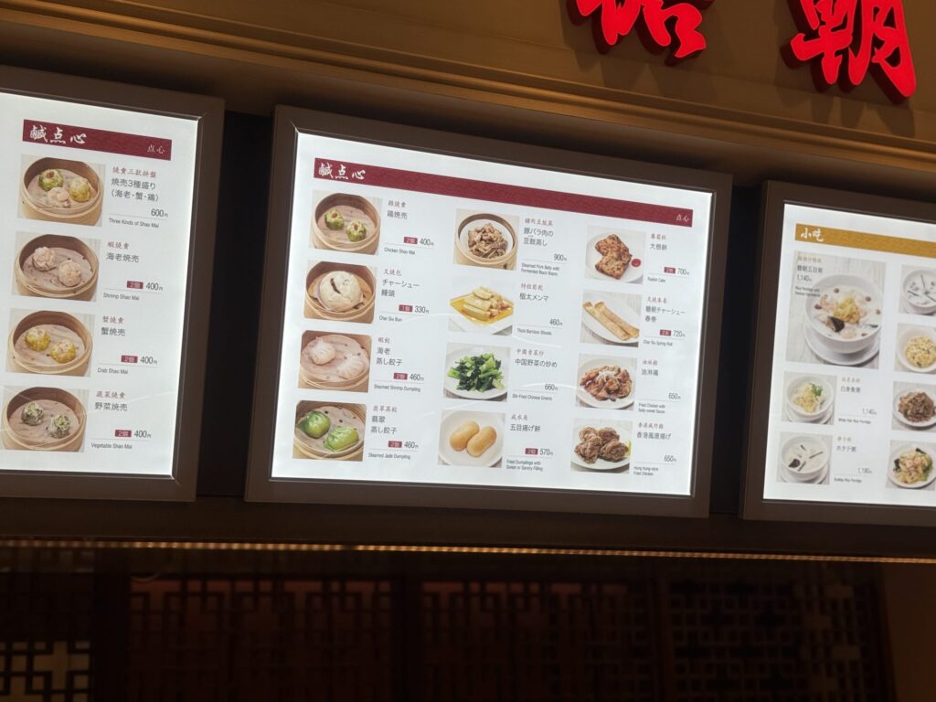 Various types of dim sum, such as shumai and gyoza, packed in a takeaway container. A sign in the background reads 'Takeaway available' in Japanese.