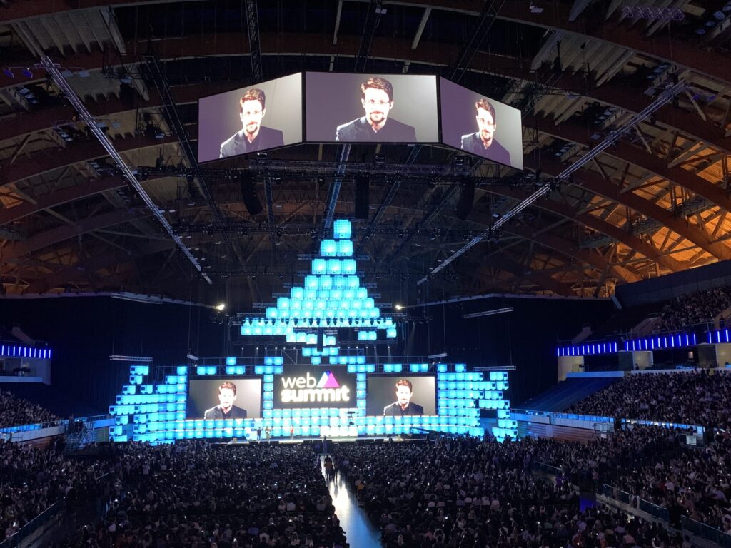 Edward Snowden appearing via video call on a large screen at an event venue, with the audience looking up at him.