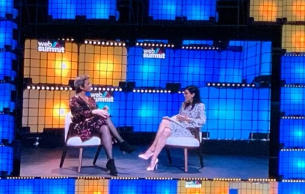 Margrethe Vestager standing on stage at Web Summit 2019, speaking into a microphone. The 'WEB SUMMIT' logo is displayed on a large screen in the background.