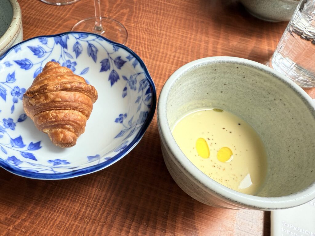 A mini croissant and a cup of cold corn soup served on a white plate on a wooden surface.
