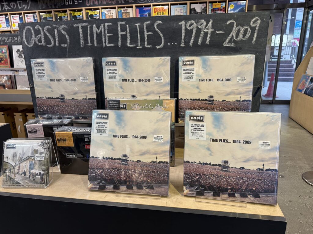 レコード店の棚に陳列されたOasisのベストアルバム「Time Flies」のLPと、「Some Might Say」の7インチシングルレコード。