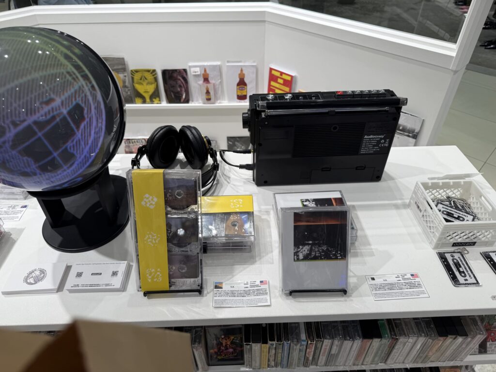 木製のテーブルの上に展示された、LAの森林火災チャリティカセットテープ。オレンジ色のカセットと、山火事を描いたジャケットが写っている。