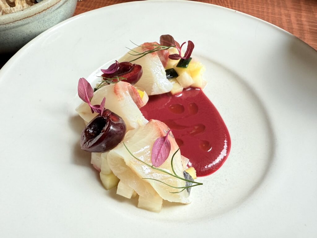 Striped jack (shima-aji) and cherry carpaccio served on a white plate. Red cherries and green herbs add a splash of color.