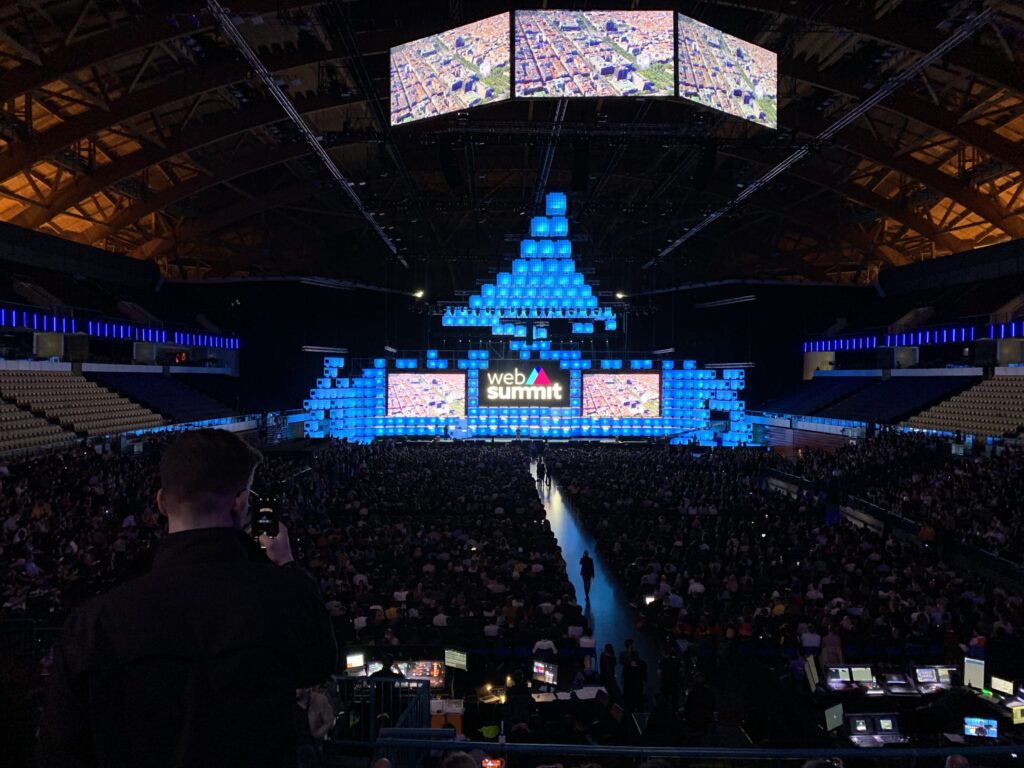 An empty conference hall before an event begins. The stage is lit with large screens, and the audience seats are vacant.