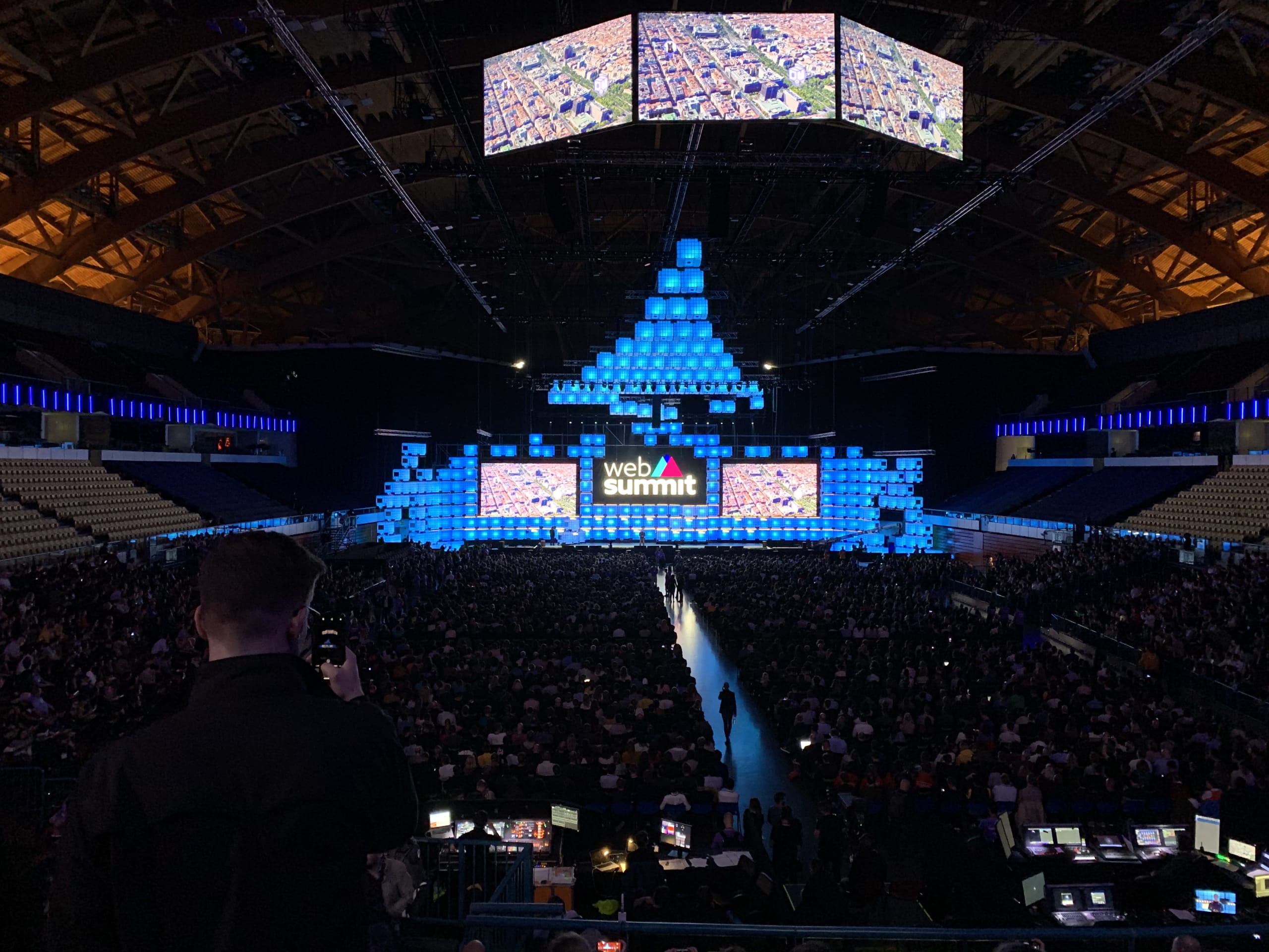 An empty conference hall before an event begins. The stage is lit with large screens, and the audience seats are vacant.