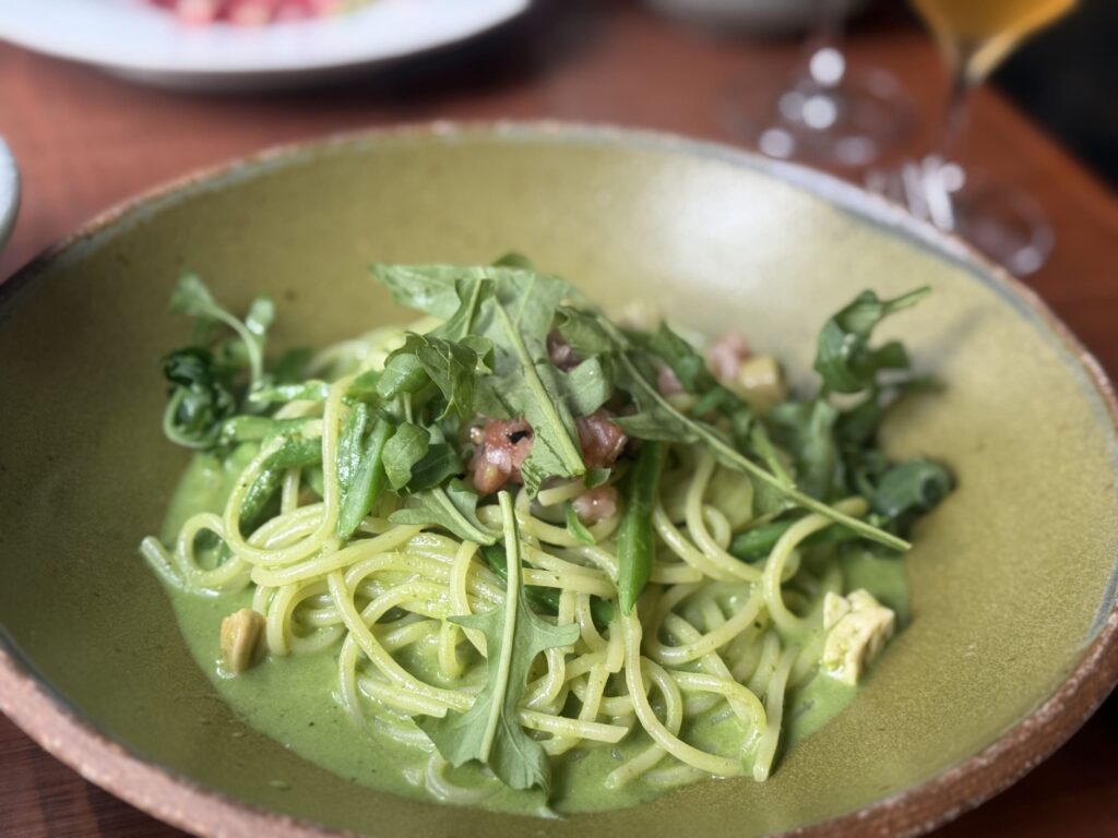 A close-up of geoduck pasta with basil sauce on a white plate. The green sauce coats the pasta, which is topped with chopped geoduck and basil leaves.
