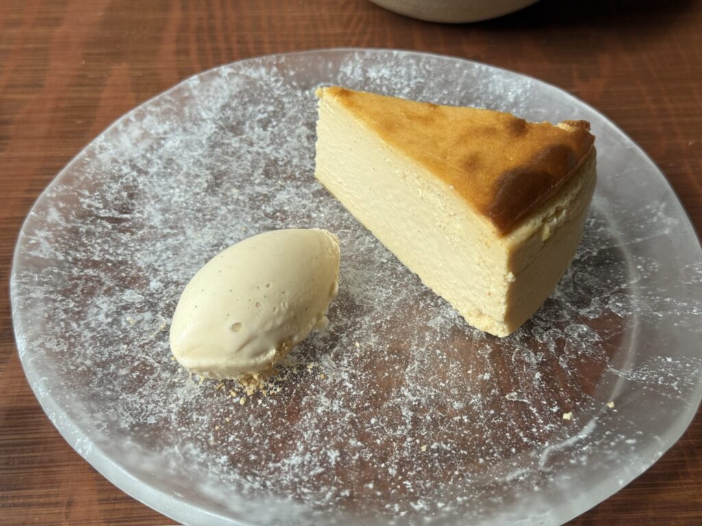 A triangular slice of baked cheesecake with a golden-brown top, served on a white plate. It is placed on a wooden table.