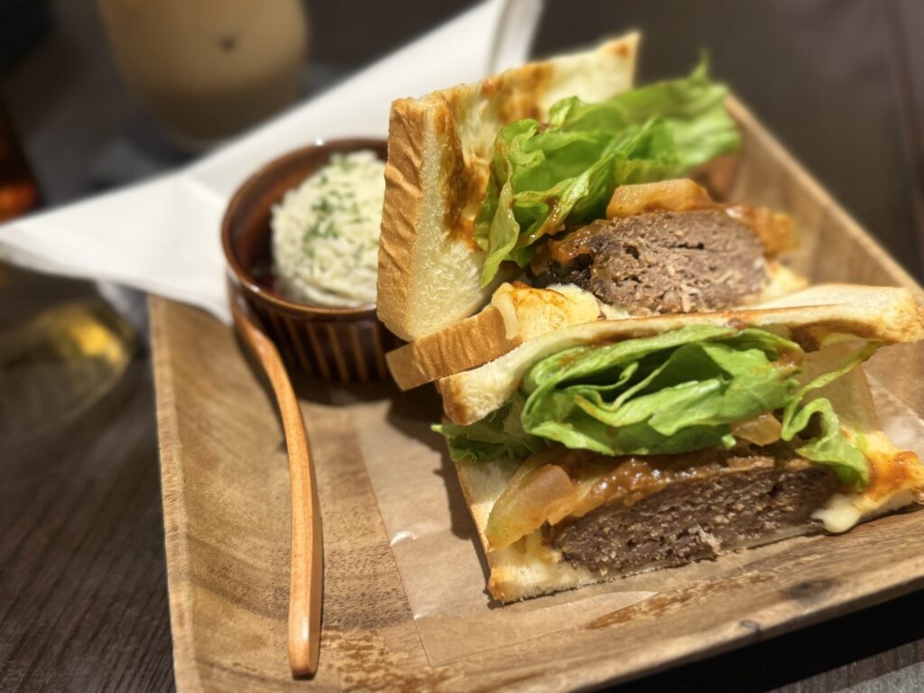 A white plate on a wooden table. A hamburg steak sandwich with demi-glace sauce and melted cheese is cut in half, served with a side of salad and french fries as a lunch set.