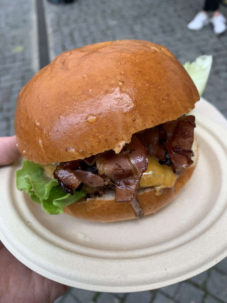A person's hand holding a paper container of french fries topped with gravy and cheese curds at an event, with other people and stalls in the background.