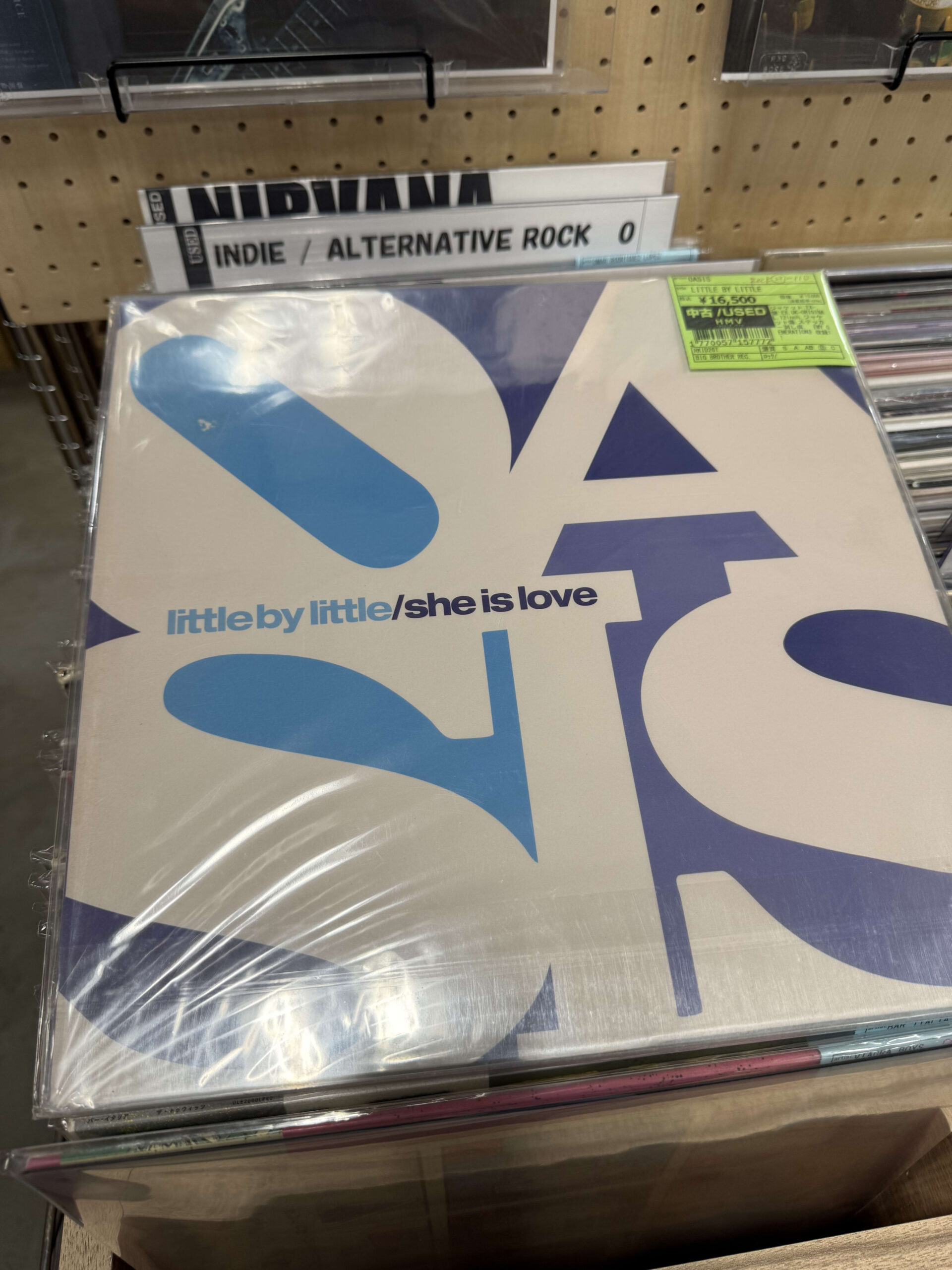 レコード店のインディー・オルタナティブの棚に置かれたOasisの12インチレコード「Little by Little / She is Love」。青いジャケットが特徴的。