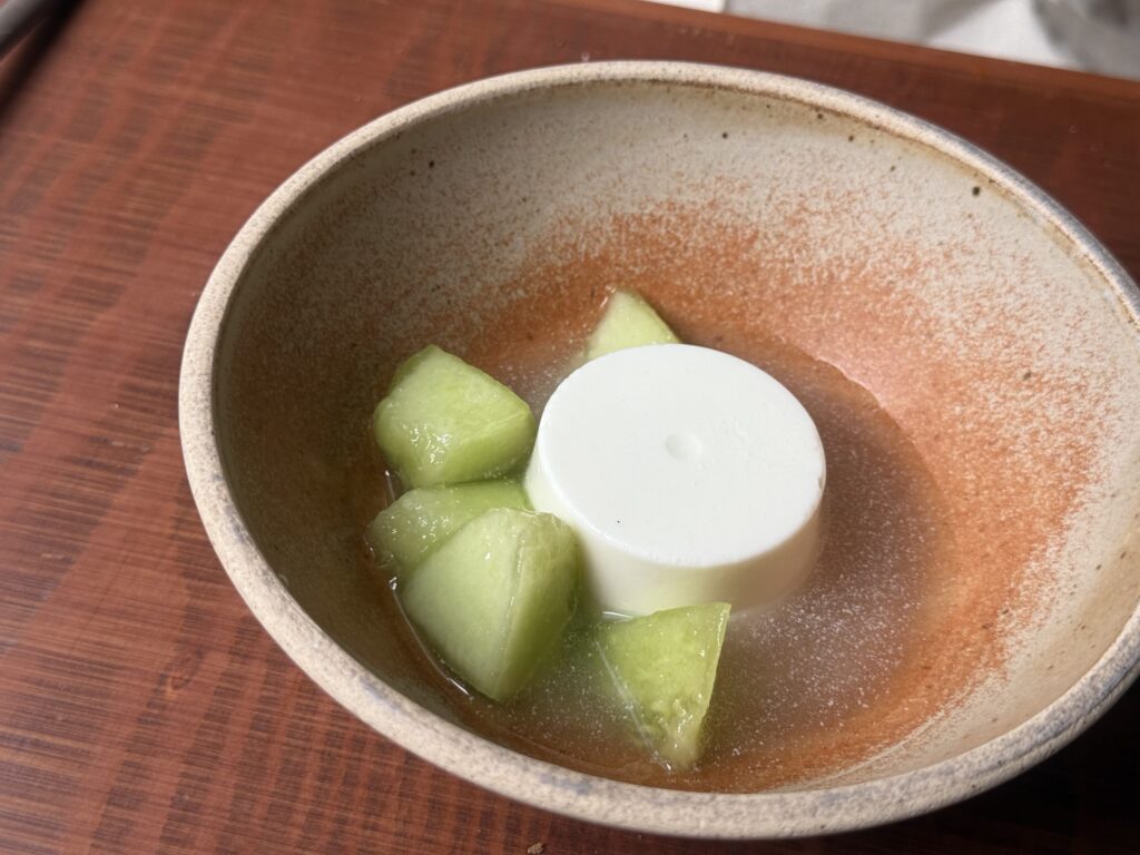 A blancmange dessert topped with basil sauce and diced melon, served on a white plate.