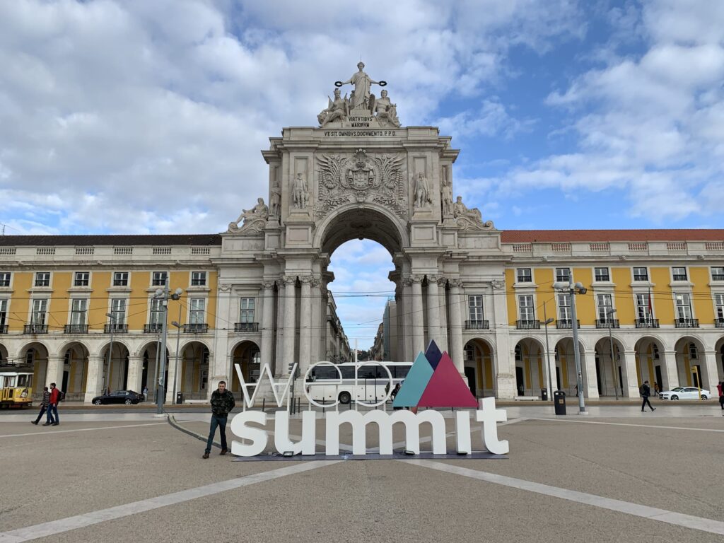 The colorful Web Summit logo monument standing in Praça do Comércio, Lisbon. The square's yellow historic buildings with arcades and a clear blue sky are visible in the background.
