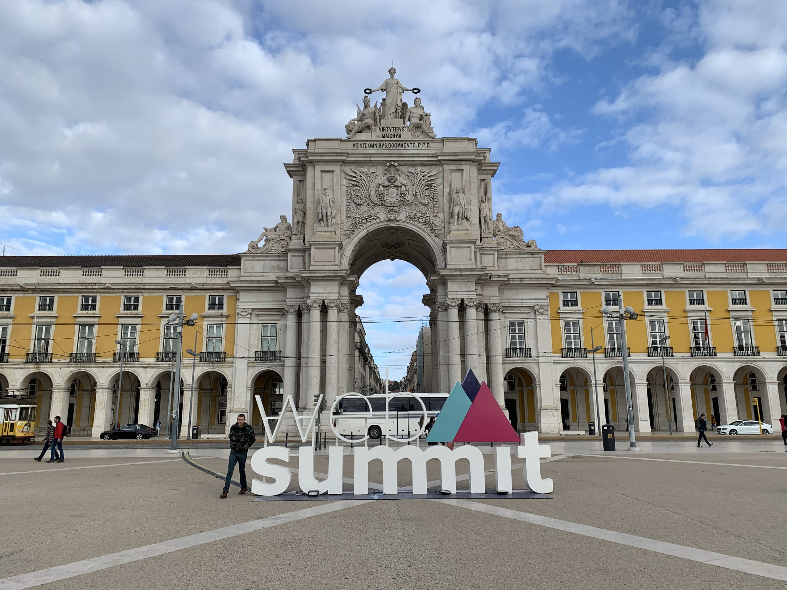 リスボンのコルメシオ広場に設置された、カラフルなWeb Summitのロゴモニュメント。背景には広場の黄色い建物とアーチ、そして青空が広がっている。