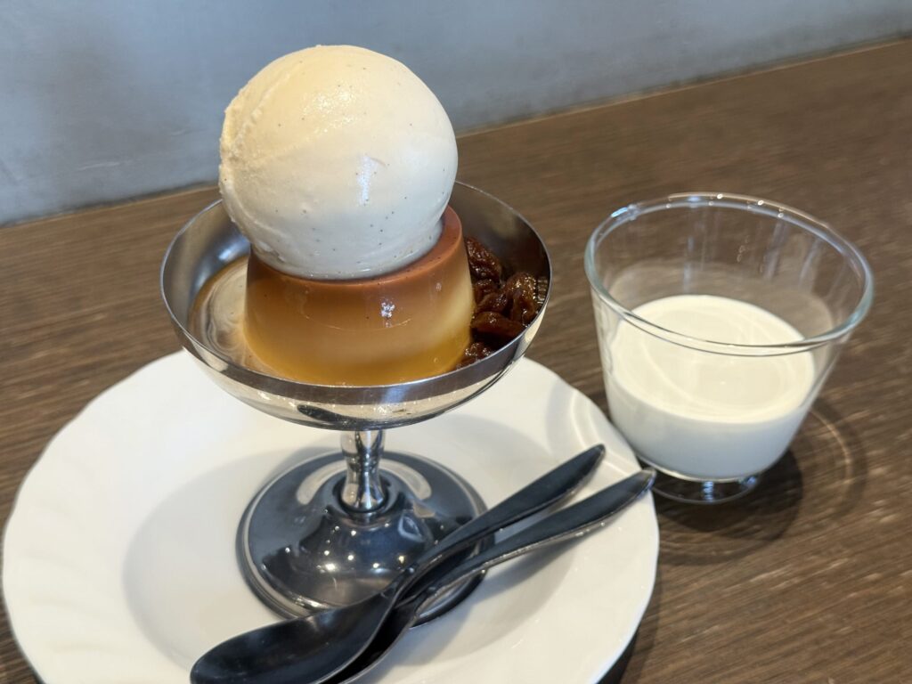 A custard pudding on a white plate. It is generously covered with a glossy caramel sauce and served with a side of whipped cream.