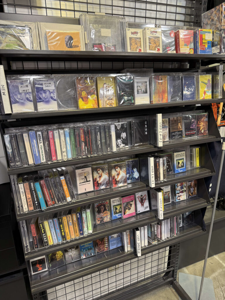 The shelves of a cassette corner in the back of a store. Many cassette tapes in their cases are packed tightly together.