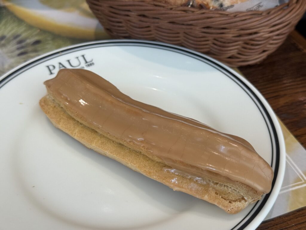 Two chocolate éclairs on a white plate at a cafe table. One is cut in half, revealing the cream filling inside.
