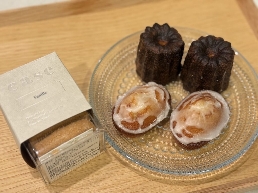 A canelé, a financier, and a vanilla cookie are arranged on a white plate, with a coffee cup in the background.