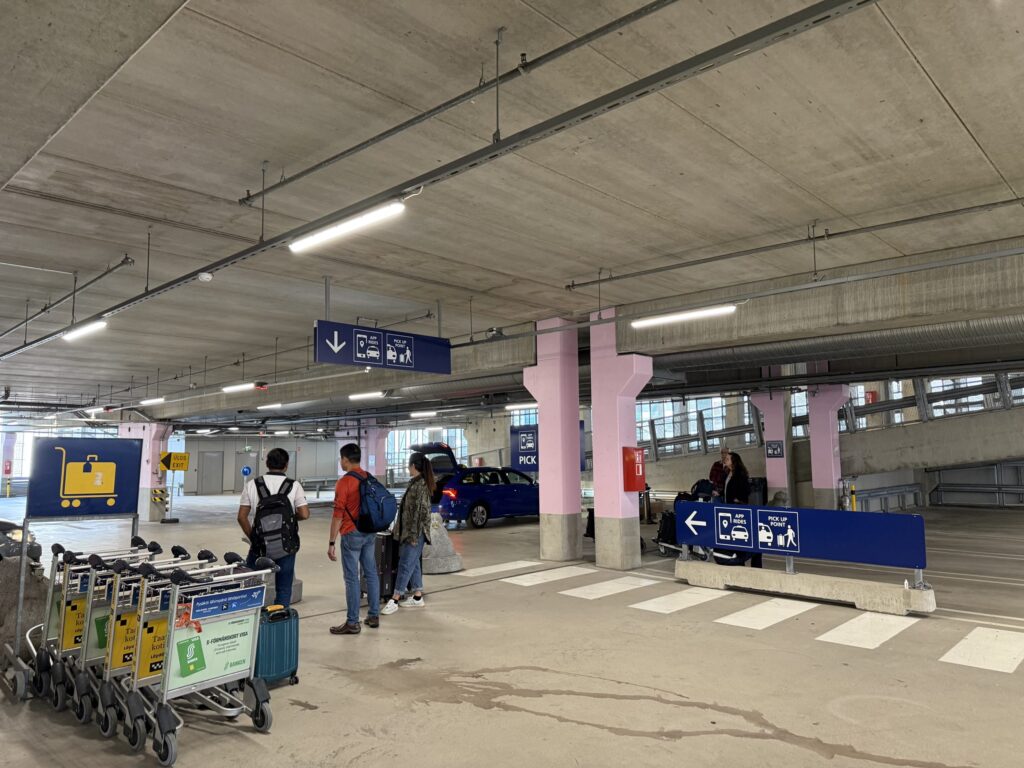 The app rides pickup point in an airport parking garage. Travelers with suitcases wait for their cars under a blue directional sign hanging from the ceiling.