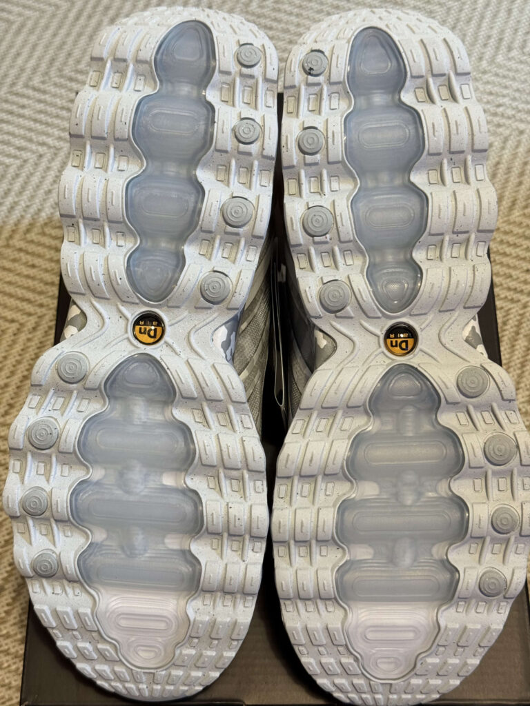 An overhead shot of the soles of a pair of Nike sneakers. The light gray speckled outsoles feature transparent air units and a yellow 'Tn AIR' logo.