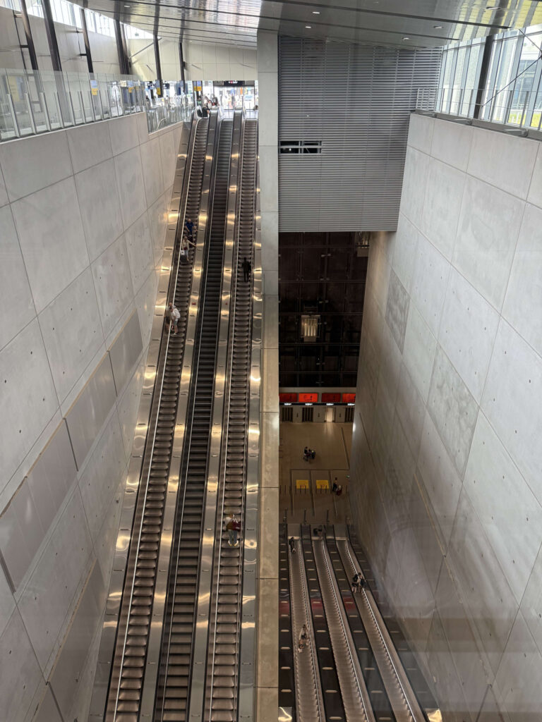 A high-angle shot of a modern subway station in Helsinki. People descend on long escalators towards the underground platform within a space enclosed by concrete walls.