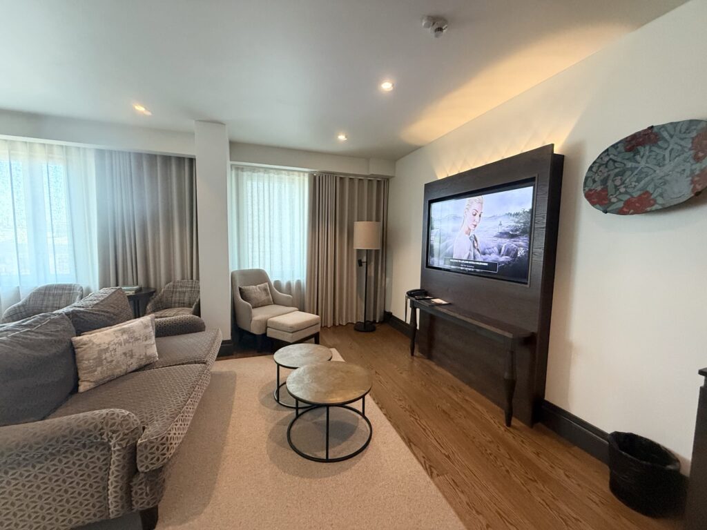 A softly lit hotel living room. It is furnished with a patterned grey sofa, a beige armchair with an ottoman, two round coffee tables, and a wall-mounted TV.