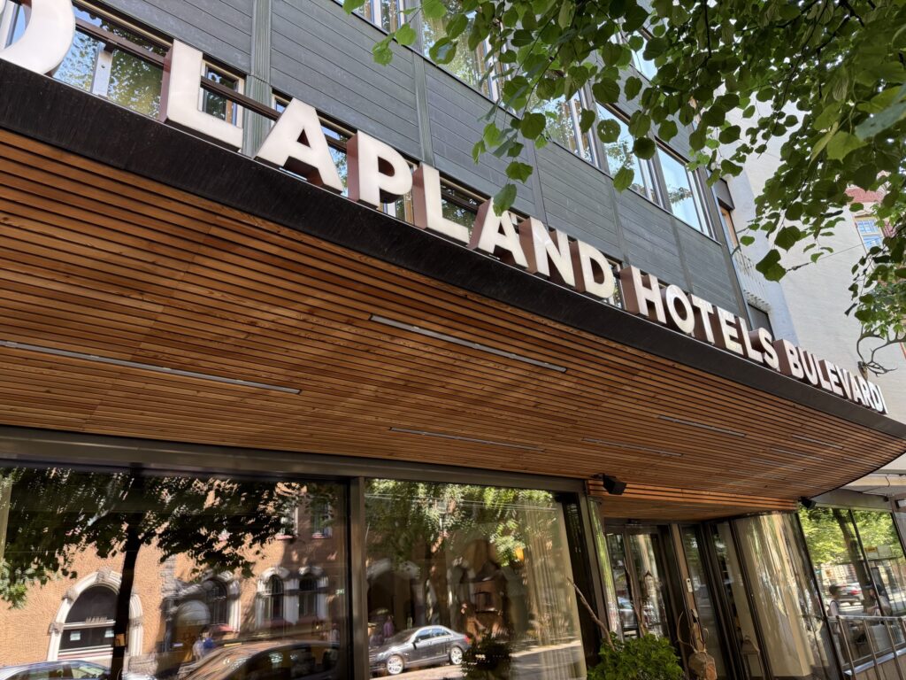 A low-angle view looking up at the entrance of Lapland Hotels Bulevardi on a sunny day. The hotel's name is in white letters above an awning with a wood-grain ceiling, and green leaves sway in the foreground.