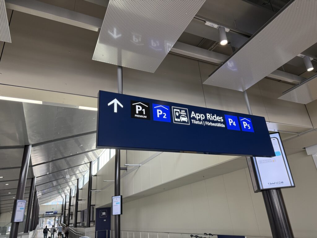 A blue overhead sign hanging from the ceiling in an airport terminal, indicating directions to parking and the 'App Rides' pickup point for ride-hailing services.