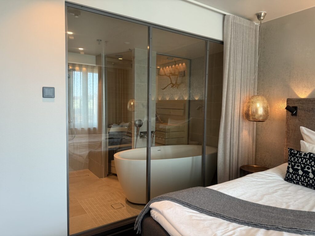 A view from the bedroom of a hotel room, showing a glass-walled bathroom with a modern white freestanding tub and a sauna in the back.