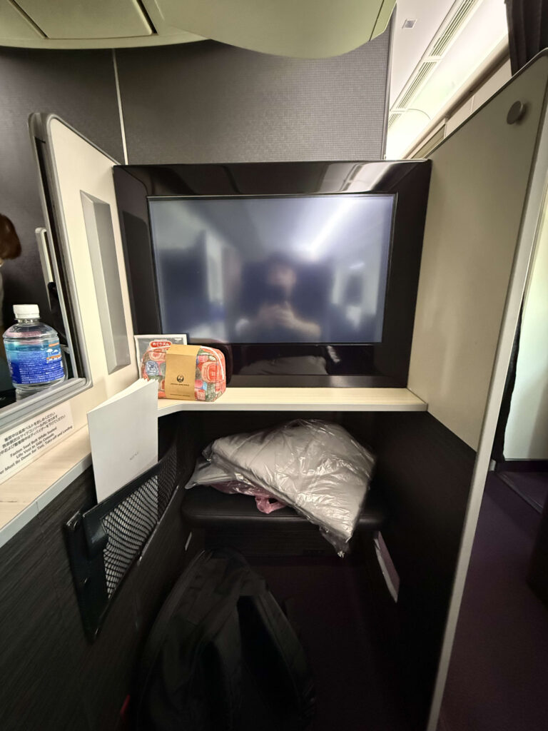 View from seat 1A of the JAL Boeing 777 Business Class 'Sky Suite', showing the personal monitor and the aircraft wing against a blue sky outside the window.