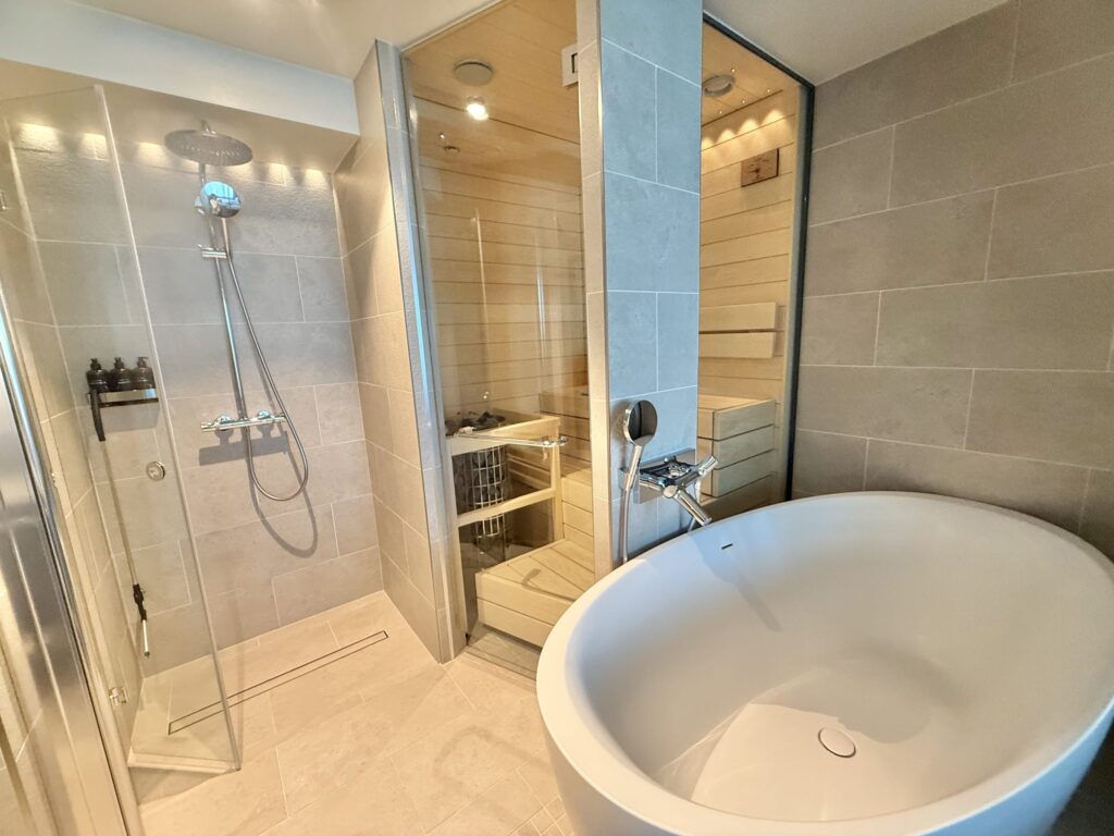 A modern bathroom featuring a walk-in glass shower, a freestanding white bathtub, and a glass-enclosed wooden home sauna.